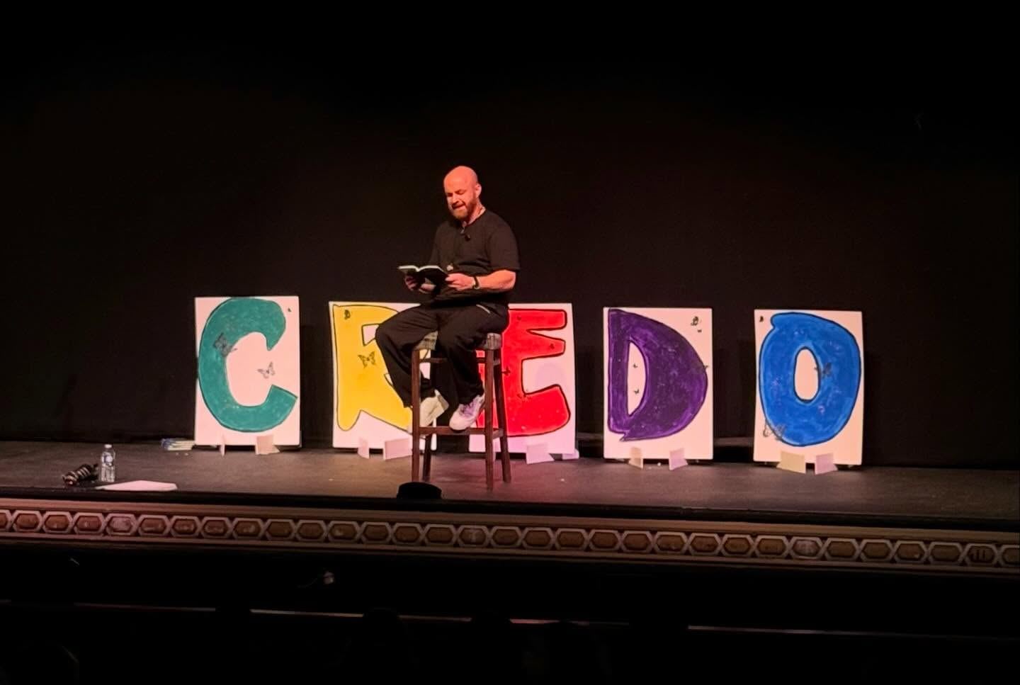 Luke Staton reading Credo on stage, seated in front of large hand-painted letters spelling CREDO.