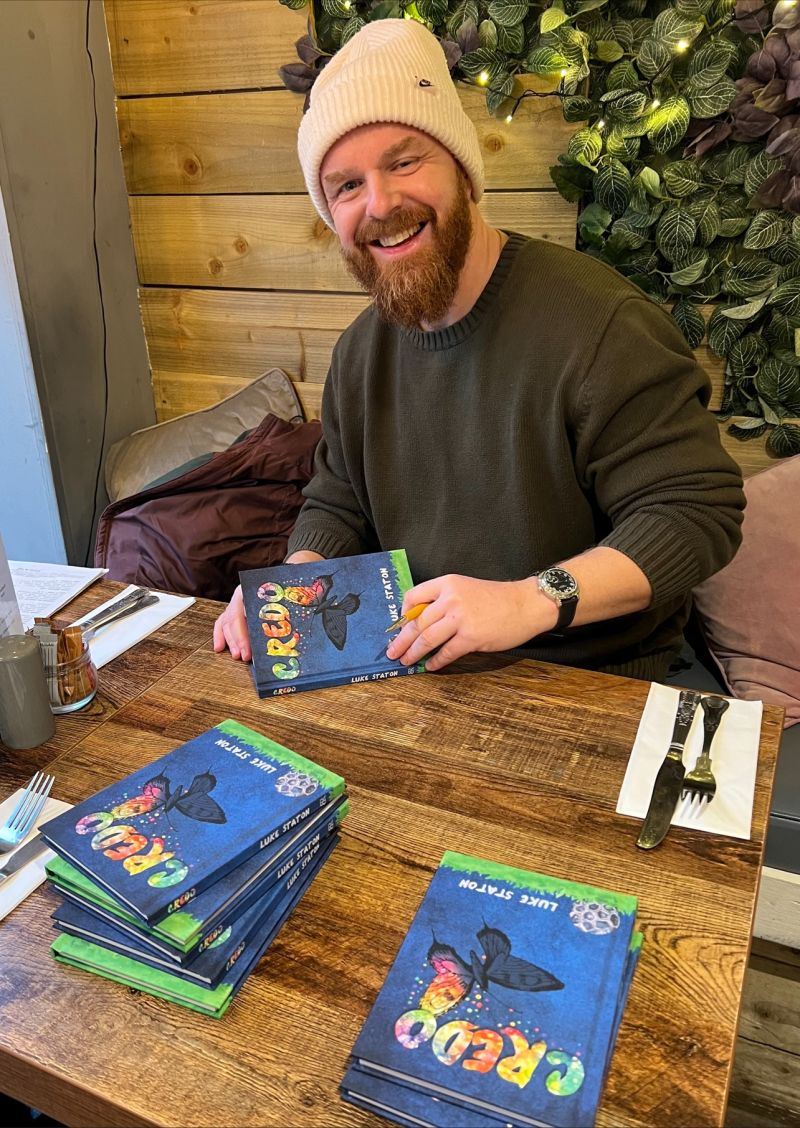 Luke Staton smiling at a wooden table, signing copies of Credo.